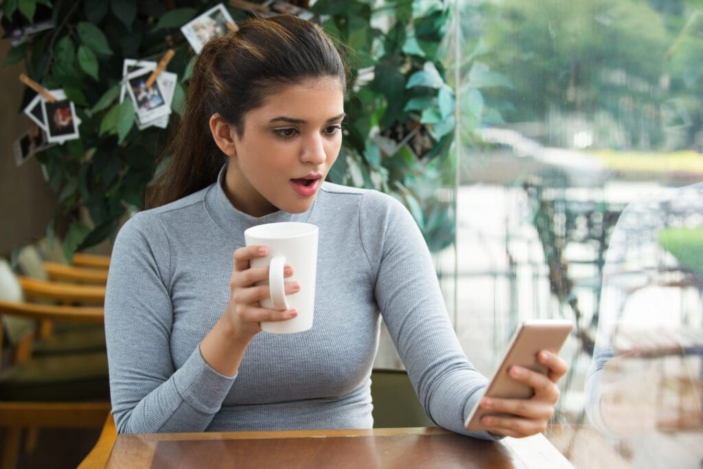 Young woman with shocked expression at cafe holding coffee mug and phone