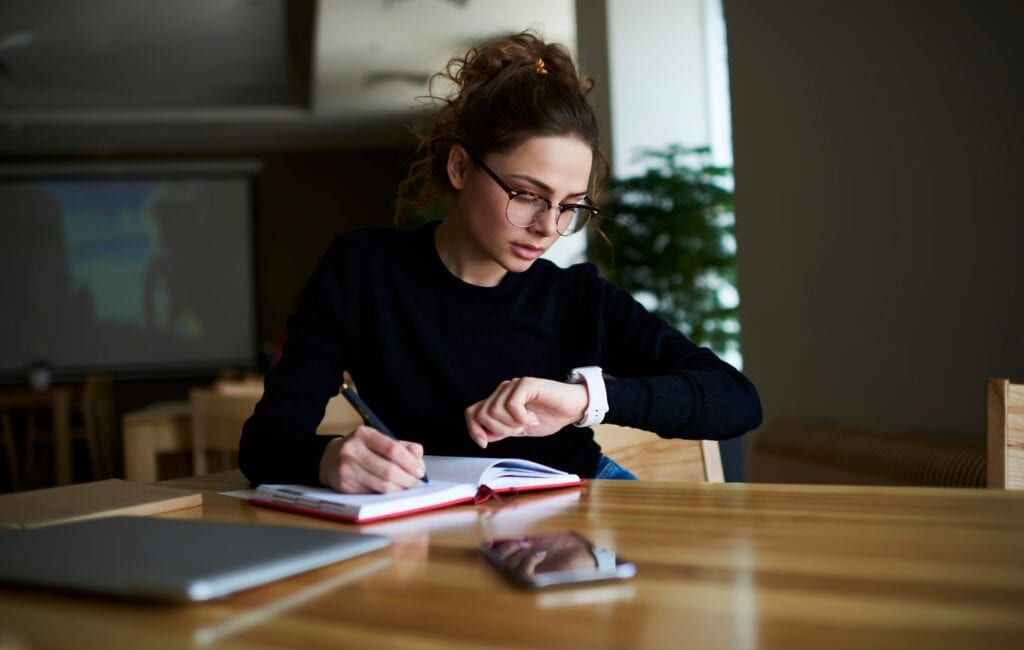 Woman wearing glasses looking at watch and writing in planner