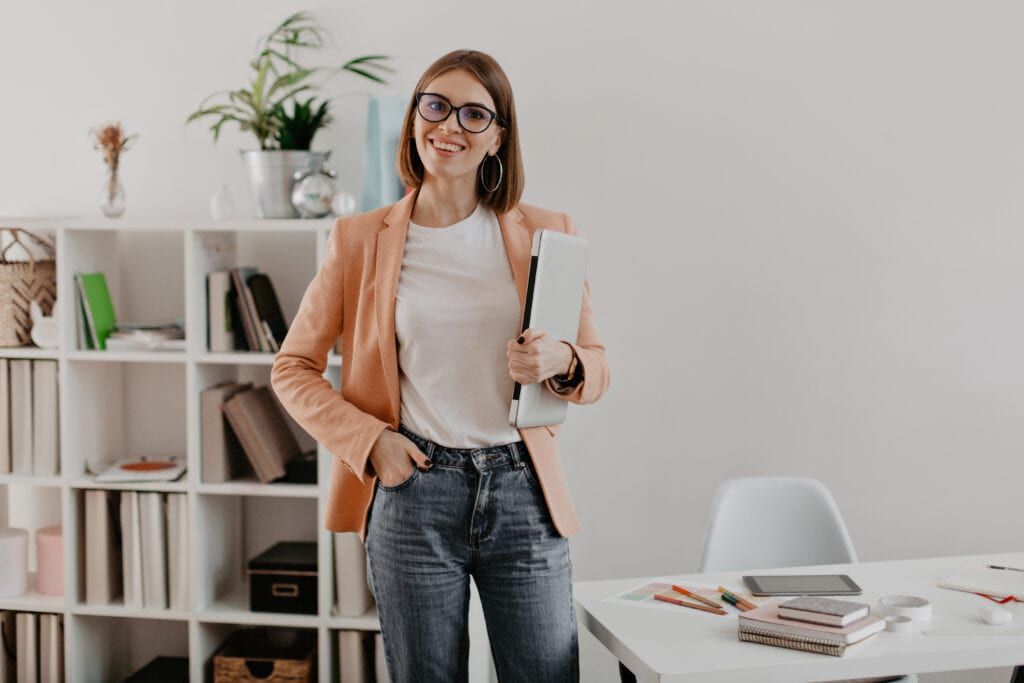 Woman wearing casual business attire holding laptop standing in home office